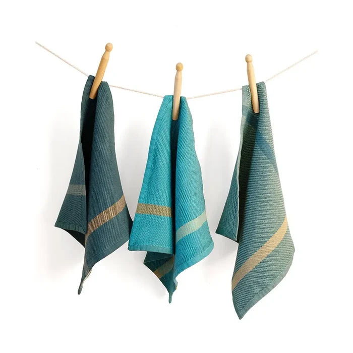 Three woven kitchen towels in teal and green tones with beige stripes, hanging from a string with wooden clothespins against a white background.