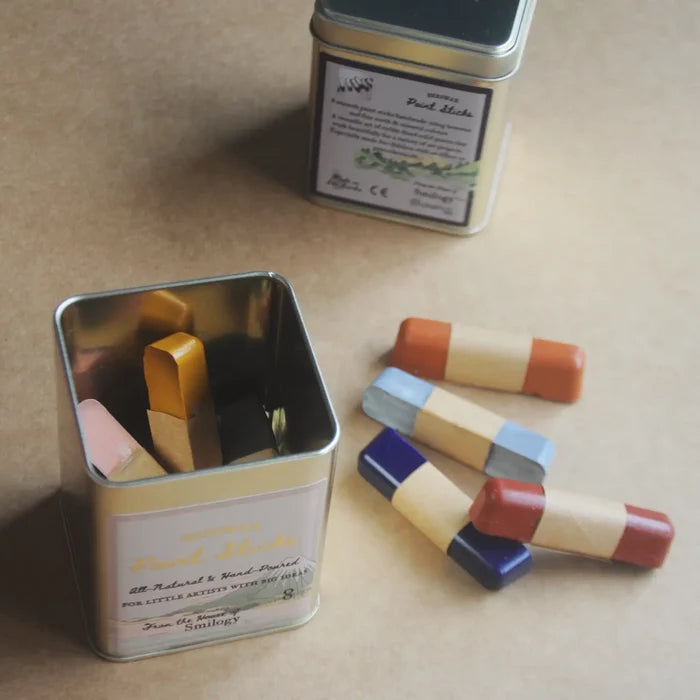 A tin of pastel sticks on a beige background, with one open tin showing the chalk inside and several loose chalk sticks arranged beside it.