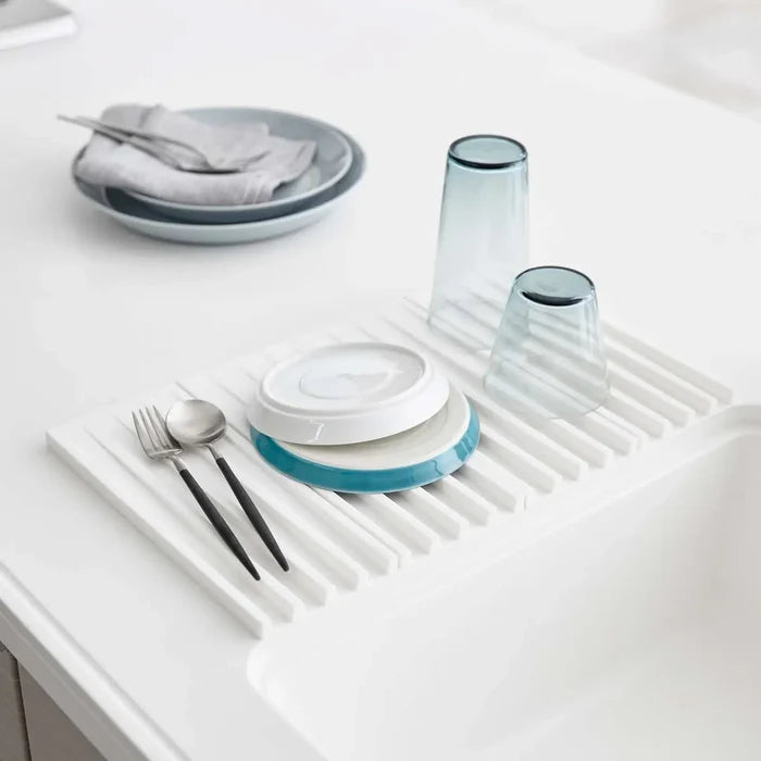 A white slotted sink drying mat shown on a white countertop, holding stacked plates, two translucent blue-gray tumblers, and utensils.