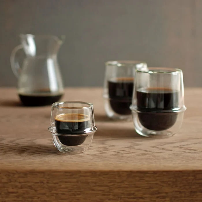 Three small glass cups with espresso on a wooden surface, with a pitcher in the background.
