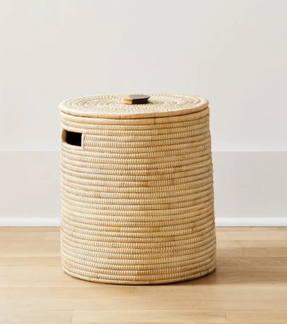 A natural woven rattan or seagrass storage basket with a matching lid and a small side cutout, standing on a light wood floor against a white wall.