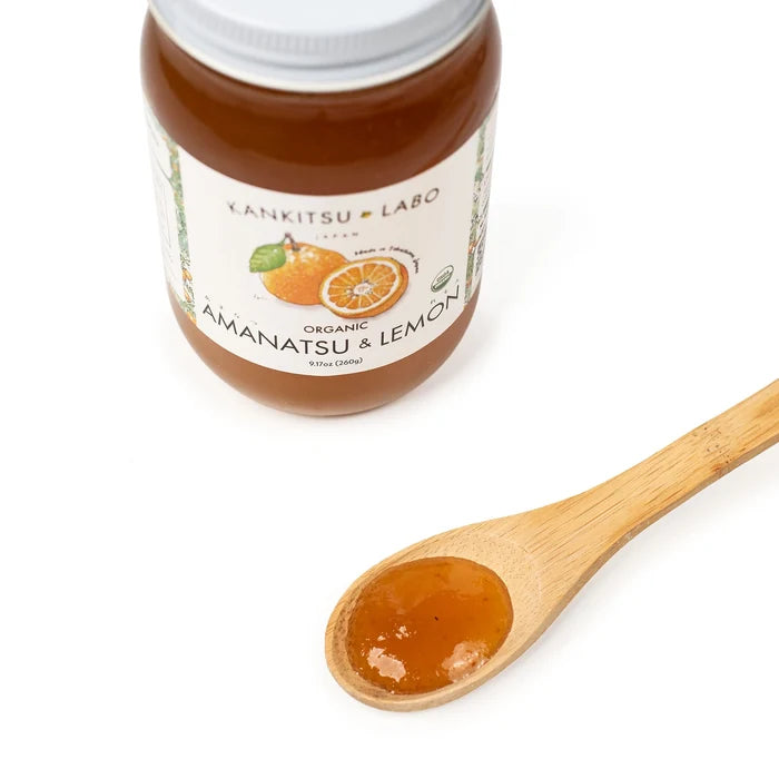 Jar of organic Amanatsu & Lemon spread with a wooden spoon containing some of the spread on a white background.