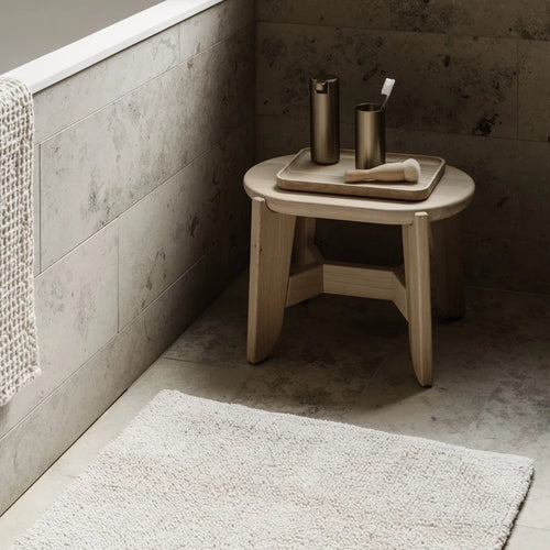 Bathroom corner with a wooden stool, bath products, and a bath mat.