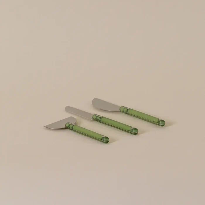 Three cheese knives with silver metal heads and translucent green handles, arranged diagonally on a plain beige background.