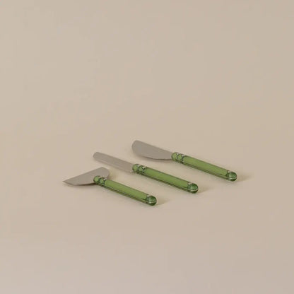 Three cheese knives with silver metal heads and translucent green handles, arranged diagonally on a plain beige background.